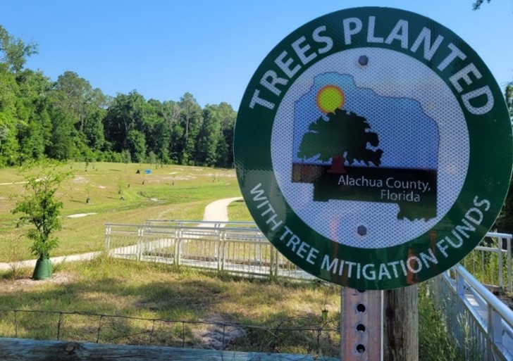 Sign reading 'Trees Planted with Tree Mitigation Funds' with an illustration of Alachua County, Florida, in the foreground. Behind the sign is a grassy open area with young trees planted throughout, a winding path, and a backdrop of dense green forest under a clear blue sky.
